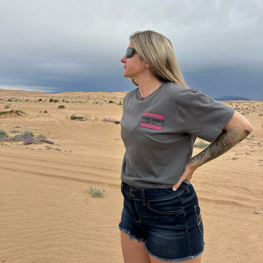 Woman standing in a desert landscape wearing a gray cropped t-shirt with a havoc honey threads logo in pink and black and denim shorts.