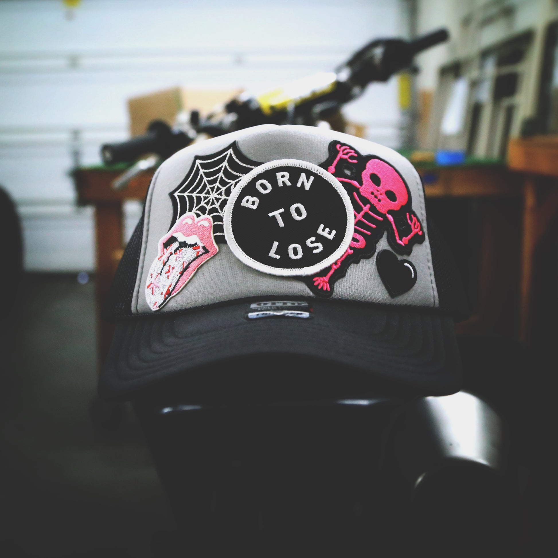 BLACK AND LIGHT GRAY TRUCKER HAT WITH 5 PATCHES. SPIDERWEB, BORN TO LOSE, LIPS WITH TONGUE, PINK SKELETON AND A SMALL HEART