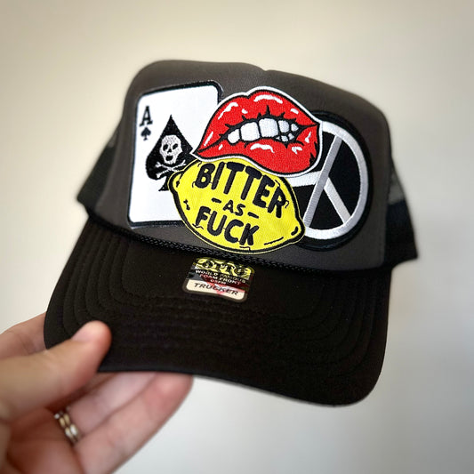 black and dark gray trucker hat with 4 patches. ace of spades, lips, peace sign and lemon that says bitter as FUCK