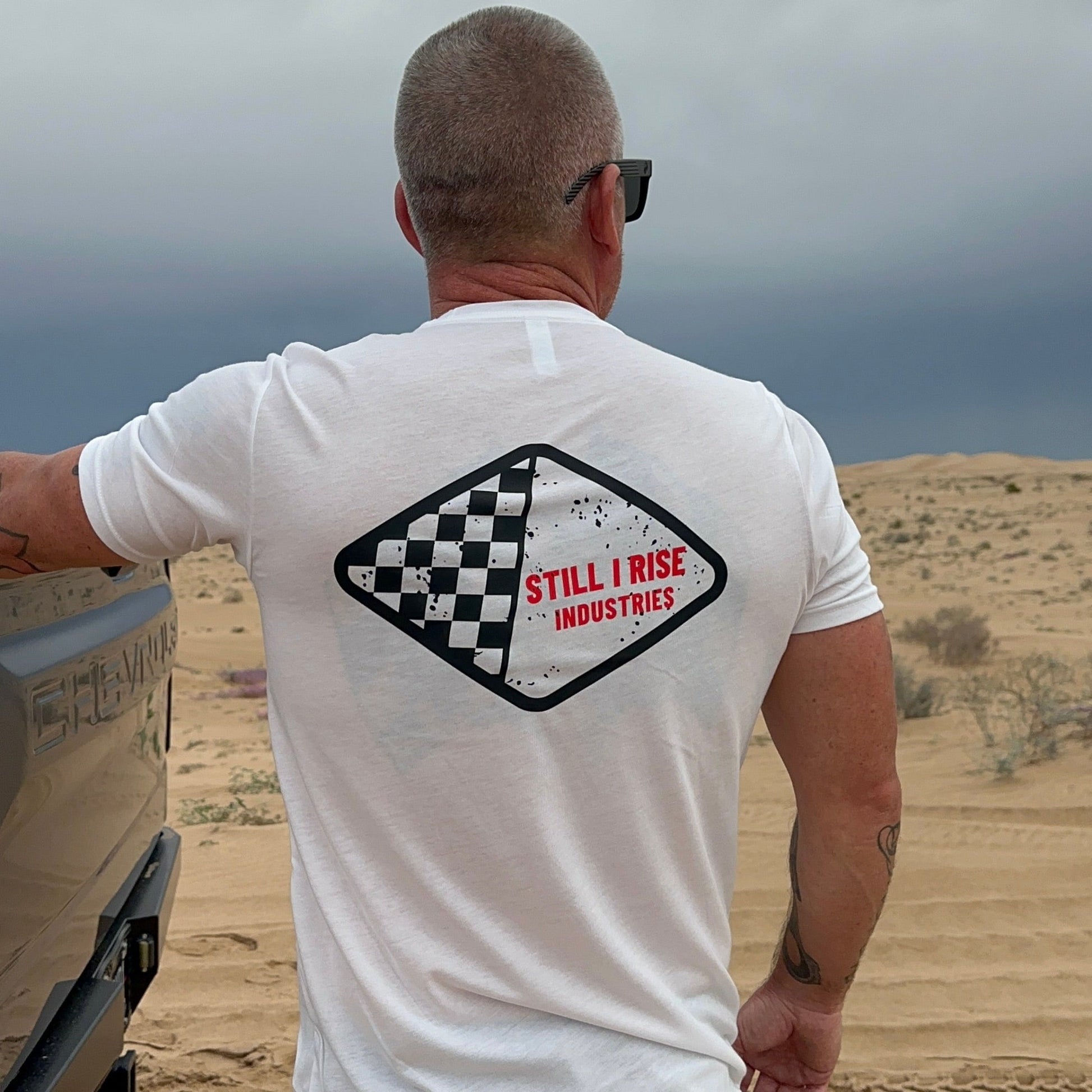 Man wearing a white t-shirt with 'Still I Rise Industries' logo in a desert setting