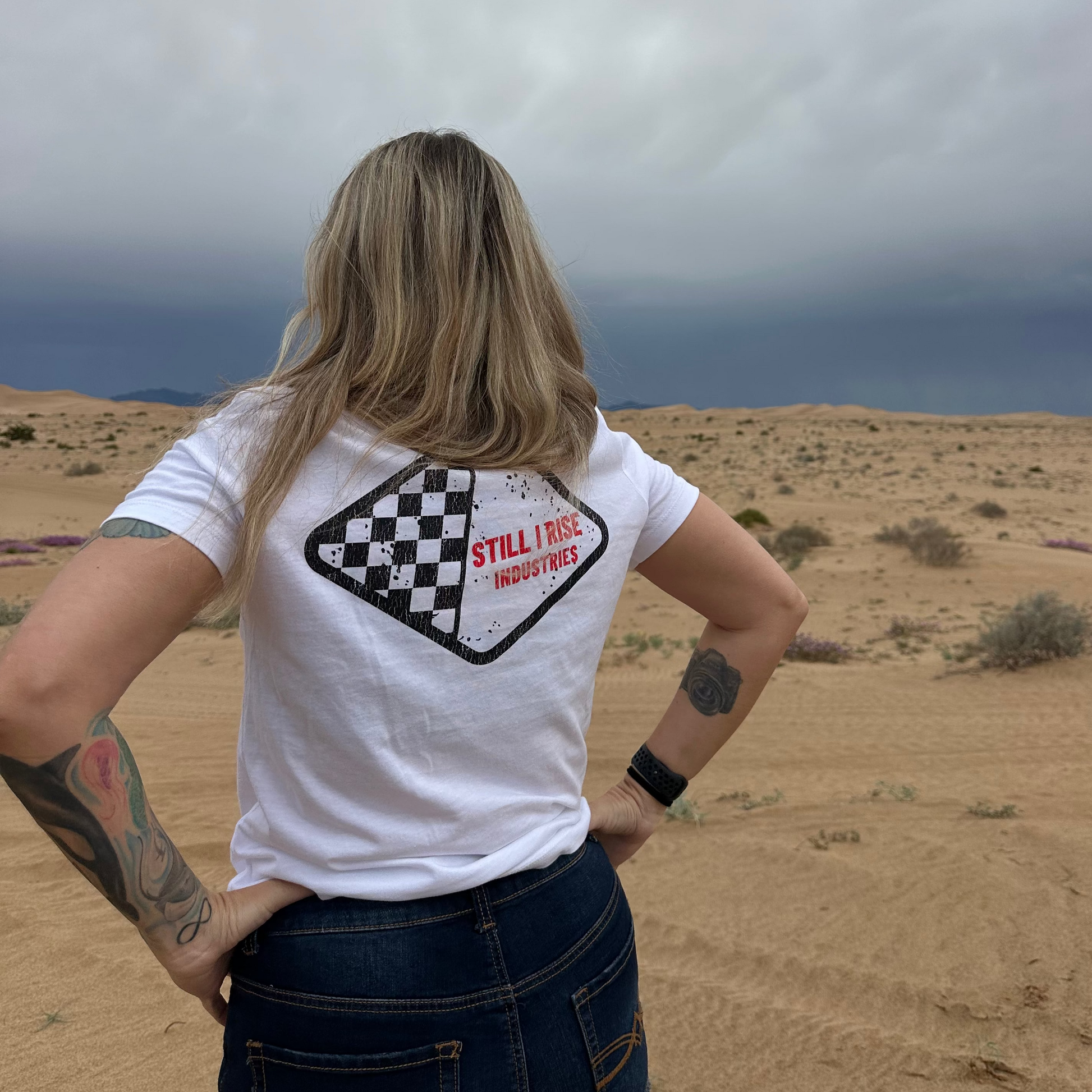 PERSON WEARING A WHITE TEE WITH A DIAMOND SHAPED STILL I RISE LOGO IN THE DESERT