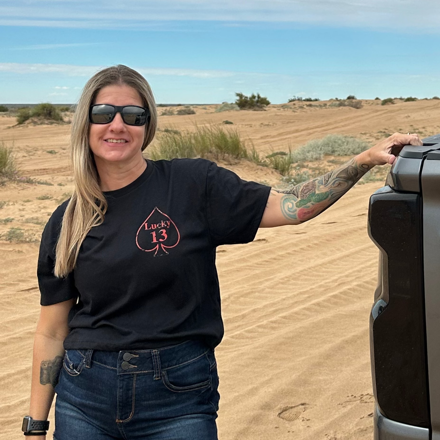 woman standing next to a truck in desert in a black cropped tee with a spade and says lucky 13 on the left breast