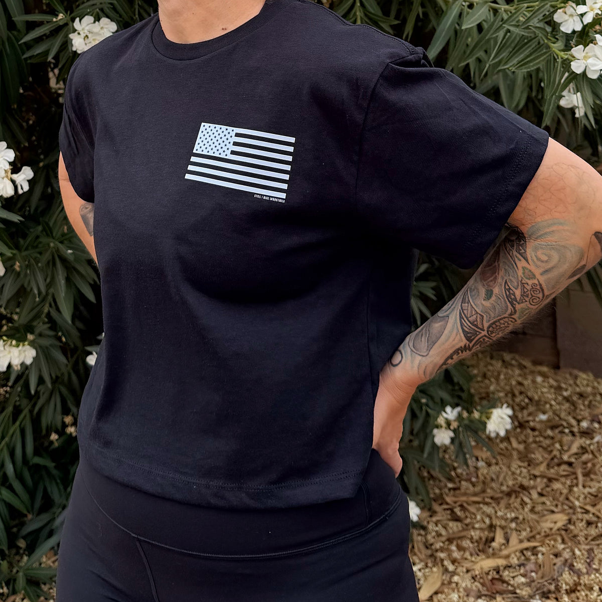Person wearing a black t-shirt with an American flag design, standing outdoors with greenery and flowers in the background.