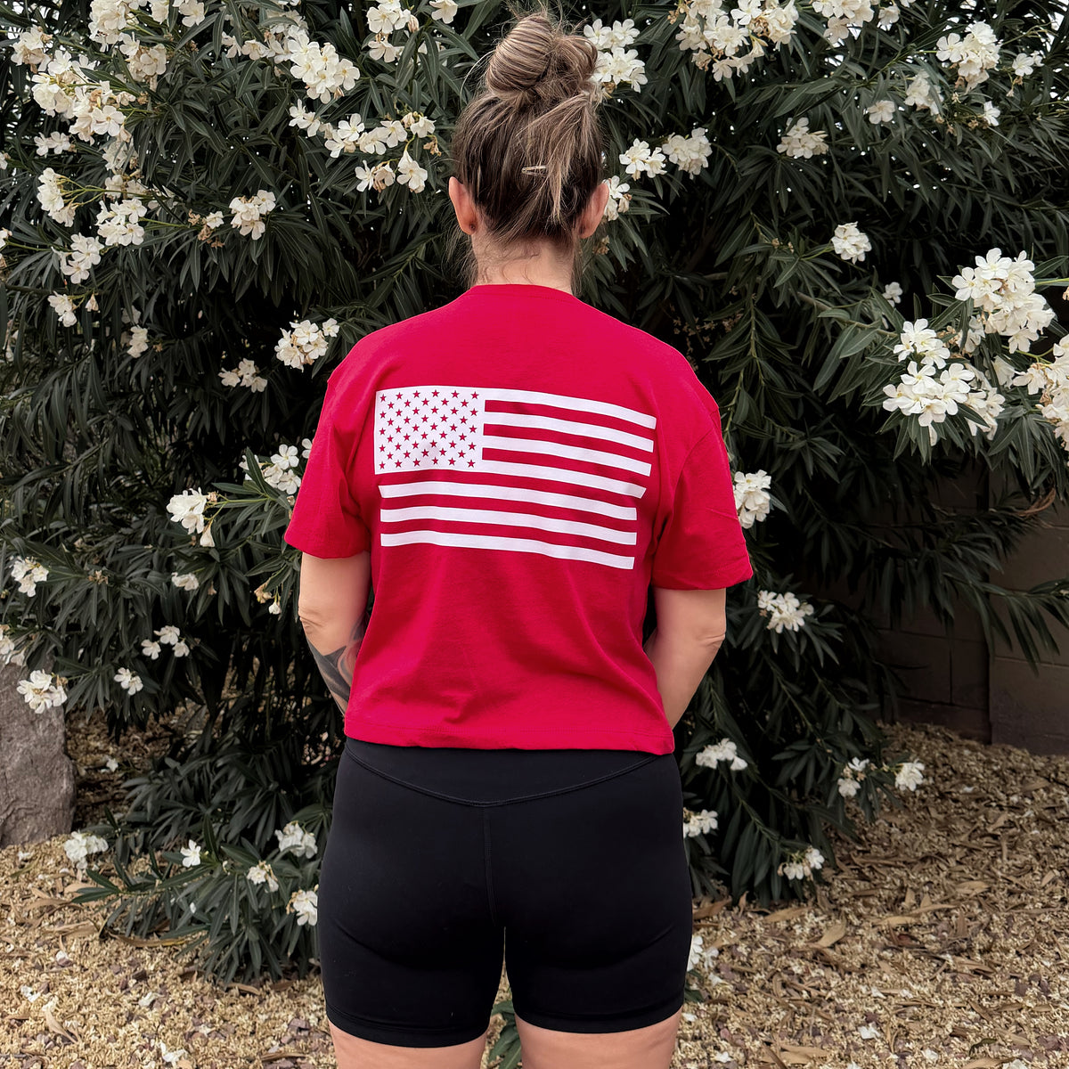 Person wearing a red t-shirt with a white flag design, standing in front of green foliage with white flowers.