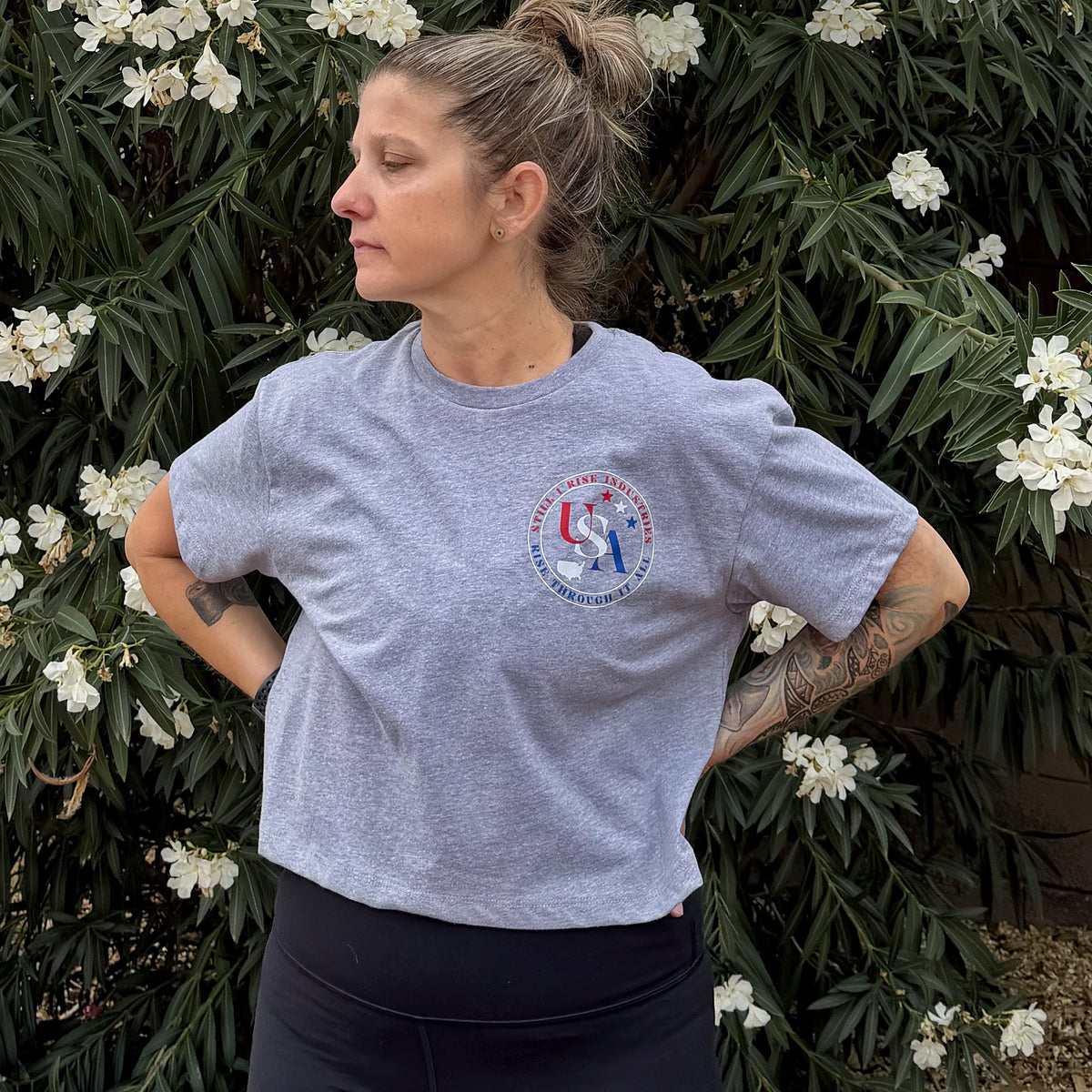 Woman wearing a gray t-shirt with USA logo, standing in front of green foliage and white flowers.