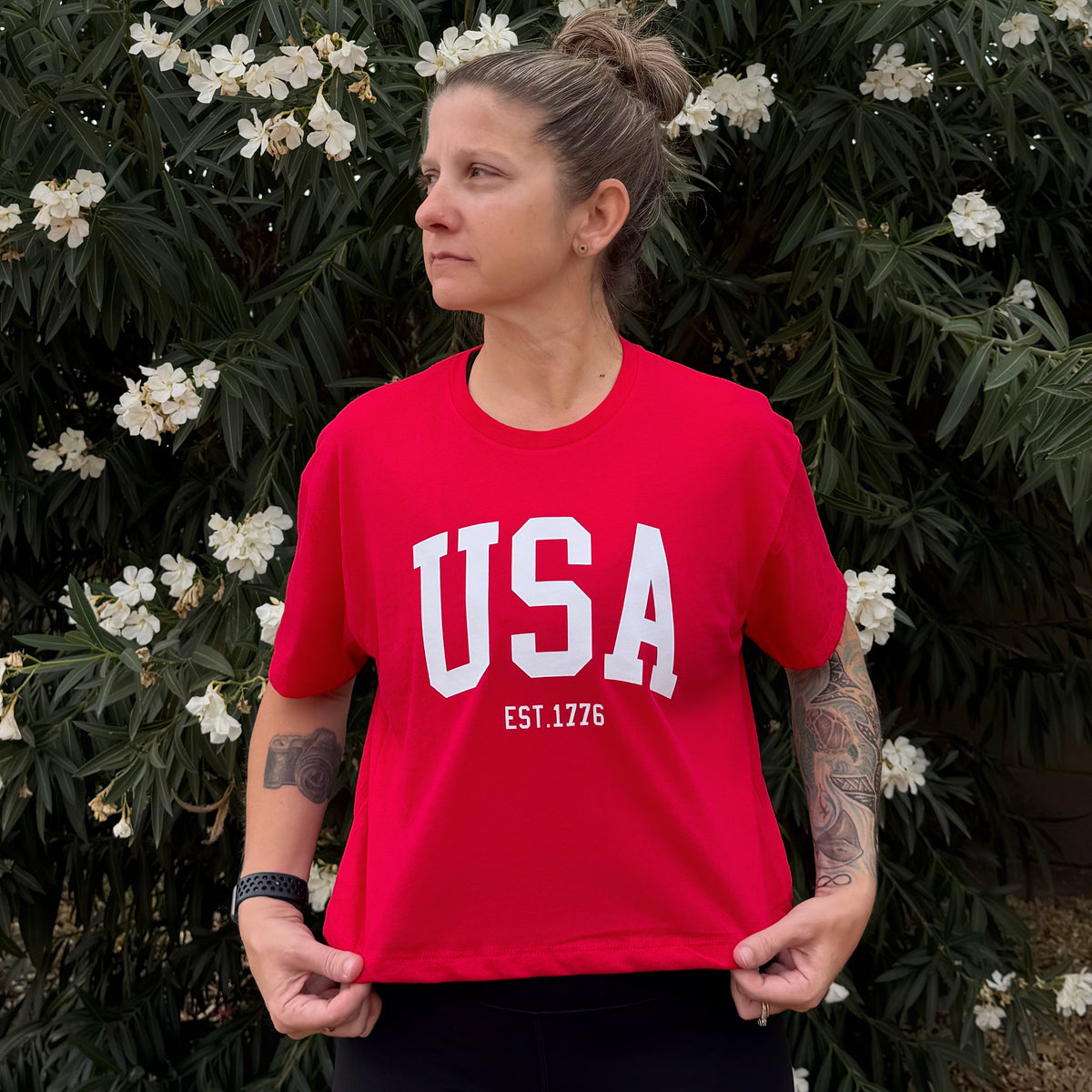 Person wearing a red 'USA' t-shirt with white text in front of green foliage and white flowers.